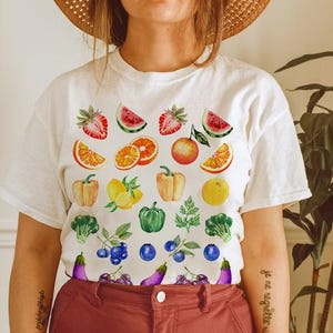 以下が含まれることがあります： 様々な果物と野菜の水彩デザインが施された白いTシャツ。デザインには、イチゴ、スイカ、オレンジ、リンゴ、パプリカ、レモン、ブロッコリー、ブルーベリー、ナスが含まれています。カジュアルな普段着です。
