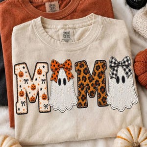 May include: Three crewneck t-shirts in black, burnt orange, and cream. The cream shirt features the word MAMA in a graphic print with pumpkins, a ghost with an orange bow, leopard print, and a ghost with a black and white plaid bow.