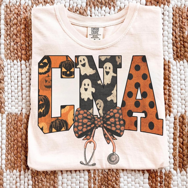 Op de afbeelding: Een licht perzikkleurig t-shirt met de letters CNA in een groot bloklettertype. De C is oranje met zwarte pompoenillustraties, de N is zwart met witte spookpatronen en de A is oranje met zwarte stippen. Een strik en stethoscoop staan eronder.