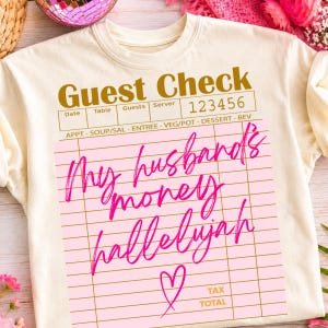 Pu&ograve; includere: Una t-shirt color crema con una grafica di un conto da ristorante stampata in oro. Il design include il testo "Guest Check" e "123456" con un riquadro rosa contenente il testo corsivo fucsia "My husband's money hallelujah" e un cuore.