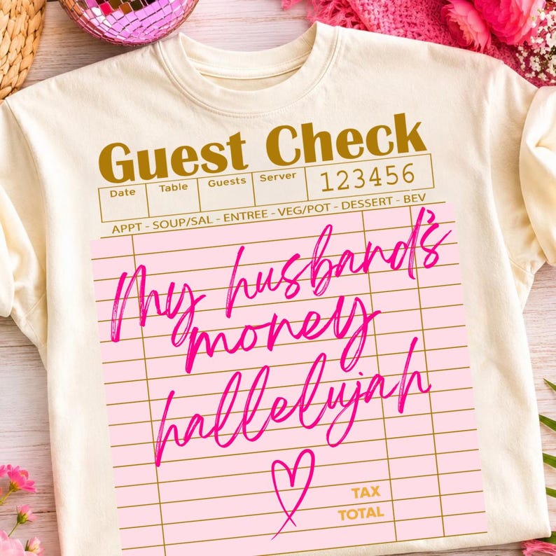 Pu&ograve; includere: Una t-shirt color crema con una grafica di un conto da ristorante stampata in oro. Il design include il testo "Guest Check" e "123456" con un riquadro rosa contenente il testo corsivo fucsia "My husband's money hallelujah" e un cuore.