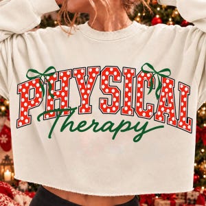Op de afbeelding: Een crèmekleurige cropped sweater met de tekst "PHYSICAL" in grote, rode blokletters met witte stippen en "Therapy" in groen cursief schrift. Het ontwerp bevat twee groene strikken aan de zijkanten van de tekst.