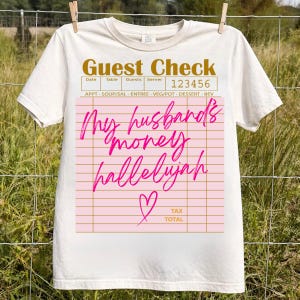 Pu&ograve; includere: Una t-shirt a maniche corte color crema con la grafica di un conto da ristorante. Il testo recita Guest Check, 123456 e My husband's money hallelujah in corsivo rosa brillante, accompagnato dal contorno di un cuore rosa e linee.