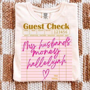 Pu&ograve; includere: Una t-shirt color crema chiaro con la grafica di un conto da ristorante. Il testo riporta Guest Check, 123456 e My husband's money hallelujah in corsivo rosa brillante, insieme a un cuore disegnato a mano e le parole Tax Total.