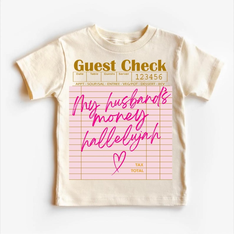 Pu&ograve; includere: Una t-shirt a maniche corte color crema con la grafica di un conto da ristorante. Il design include la scritta "Guest Check" in oro e il testo scritto a mano in rosa "My husband's money hallelujah" con un piccolo simbolo a cuore.