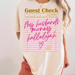 Pu&ograve; includere: Una t-shirt color crema con la grafica di un conto da ristorante. Il testo recita "Guest Check" in alto, "123456" nell'angolo e "My husband's money hallelujah" in corsivo rosa brillante, accompagnato da una piccola icona a cuore.