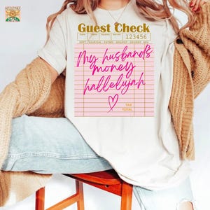 Pu&ograve; includere: Una t-shirt color crema con la grafica di un conto da ristorante. Il testo recita Guest Check 123456, My husband's money hallelujah e Tax Total, con un'icona a forma di cuore rosa. La t-shirt &egrave; indossata con un cardigan in maglia beige e jeans blu.