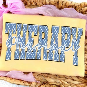 May include: A pale yellow t-shirt featuring the word THERAPY in large, block-style blue letters with a white polka dot pattern. The word physical is written across the center in a white, cursive script font with a thin blue outline.