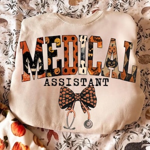 May include: A cream-coloured crewneck sweatshirt featuring the text MEDICAL ASSISTANT in large block letters with orange and black Halloween patterns, including pumpkins and ghosts. Below the text is a bow graphic with a stethoscope attached.
