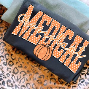 Puede incluir: Una camiseta gris oscuro con el texto "MEDICAL ASSISTANT" en letras grandes de bloque naranja con puntos blancos. Una calabaza naranja con puntos blancos está colocada sobre las letras. La camiseta está doblada sobre una superficie.