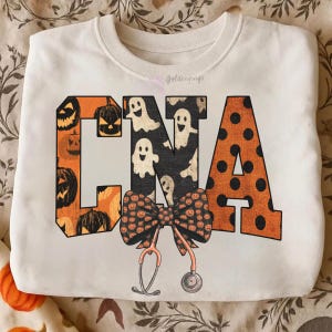 Op de afbeelding: Een cr&egrave;mekleurig sweatshirt met ronde hals en de letters CNA in een groot bloklettertype. De C is oranje met zwarte pompoenillustraties, de N is zwart met witte spookafbeeldingen en de A is oranje met zwarte stippen. Daaronder bevinden zich een strik en een stethoscoop.