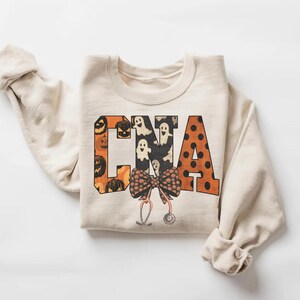 Op de afbeelding: Een cr&egrave;mekleurig sweatshirt met ronde hals en de letters CNA in een groot bloklettertype. De letters zijn gevuld met patronen van oranje pompoenen, witte spoken en zwarte stippen. Onder de letters zijn een strik en een stethoscoop afgebeeld.