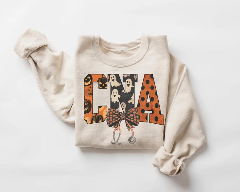 Op de afbeelding: Een cr&egrave;mekleurig sweatshirt met ronde hals en de letters CNA in een groot bloklettertype. De letters zijn gevuld met patronen van oranje pompoenen, witte spoken en zwarte stippen. Onder de letters zijn een strik en een stethoscoop afgebeeld.