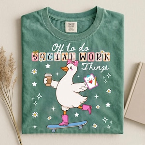 May include: A sage green t-shirt featuring a graphic of a white duck wearing a pink bow and pink boots, riding a skateboard while holding a coffee cup and a clipboard. The text reads "Off to do SOCIAL WORK Things" with small white daisy and star accents.
