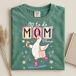 Może przedstawiać: Szałwiowo-zielony t-shirt z grafiką białej gęsi na deskorolce, w różowych butach, trzymającej kubek kawy i torbę z napisem Cool Mom. Tekst brzmi Off to do MOM Things, w otoczeniu gwiazdek i stokrotek.