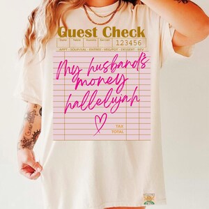 Pu&ograve; includere: Una t-shirt a maniche corte color crema con la grafica di un conto da ristorante. Il testo recita Guest Check, 123456 e My husband's money hallelujah in corsivo rosa brillante, accompagnato da una piccola icona a cuore e linee.