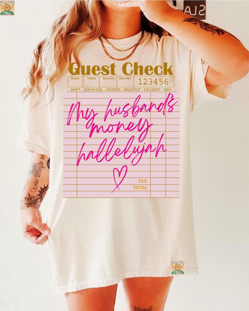 Pu&ograve; includere: Una t-shirt a maniche corte color crema con la grafica di un conto da ristorante. Il testo recita Guest Check, 123456 e My husband's money hallelujah in corsivo rosa brillante, accompagnato da una piccola icona a cuore e linee.