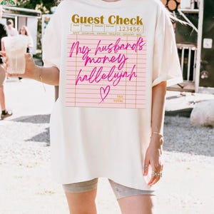 Pu&ograve; includere: Una t-shirt a maniche corte color bianco sporco con una grafica di un conto da ristorante stampata in oro e rosa. Il testo recita Guest Check, 123456 e My husband's money hallelujah in corsivo, accompagnato da una piccola icona a cuore rosa.