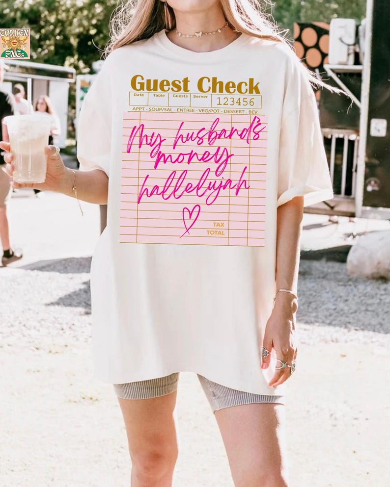 Pu&ograve; includere: Una t-shirt a maniche corte color bianco sporco con una grafica di un conto da ristorante stampata in oro e rosa. Il testo recita Guest Check, 123456 e My husband's money hallelujah in corsivo, accompagnato da una piccola icona a cuore rosa.