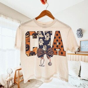 Op de afbeelding: Een cr&egrave;mekleurig T-shirt met korte mouwen en de letters CNA in een groot bloklettertype. De C is oranje met pompoenafbeeldingen, de N is donkerblauw met witte spoken en de A is oranje met zwarte stippen. Daaronder staan een strik en een stethoscoop.
