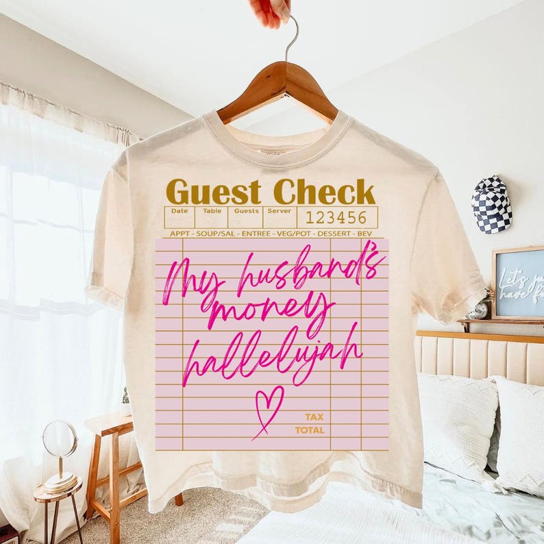 Pu&ograve; includere: Una t-shirt color crema con la grafica di un conto da ristorante. Il testo "Guest Check" appare in alto, "123456" nell'angolo e "My husband's money hallelujah" in corsivo rosa brillante, con un cuore rosa e "TAX TOTAL" in basso.