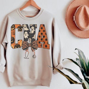 Op de afbeelding: Een lichtbeige sweatshirt met lange mouwen en de letters CNA in een groot bloklettertype. De letters zijn gevuld met patronen van oranje pompoenen, witte spoken en oranje stippen. Een strik en een stethoscoop zijn in het midden eronder geplaatst.