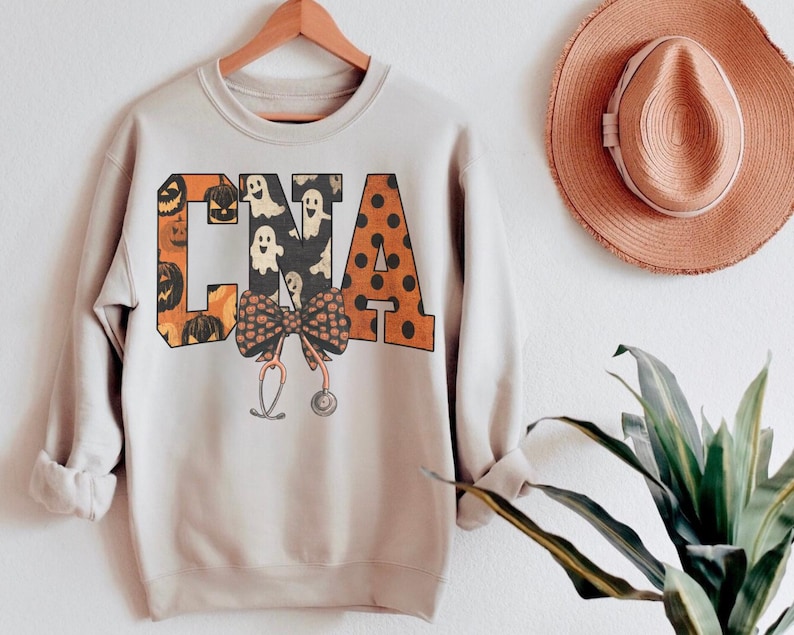 Op de afbeelding: Een lichtbeige sweatshirt met lange mouwen en de letters CNA in een groot bloklettertype. De letters zijn gevuld met patronen van oranje pompoenen, witte spoken en oranje stippen. Een strik en een stethoscoop zijn in het midden eronder geplaatst.