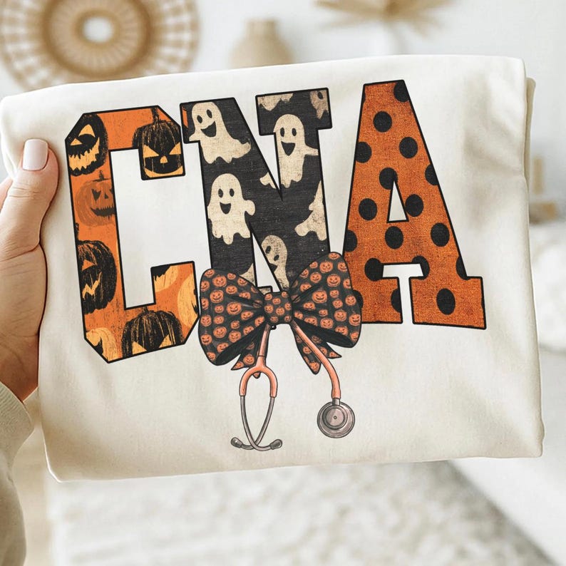Op de afbeelding: Een cr&egrave;mekleurig t-shirt met de letters CNA in een groot bloklettertype. De C is oranje met zwarte pompoenen, de N is zwart met witte geesten en de A is oranje met zwarte stippen. Daaronder bevinden zich een strik en een stethoscoop.
