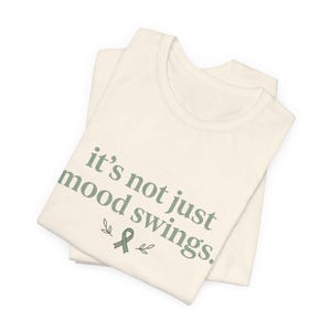 Könnte beinhalten: Cremefarbenes T-Shirt mit dem Text "it's not just mood swings." in einer gedeckten grünen Schrift. Das Design beinhaltet ein grünes Band und Blätter. Das Shirt ist gefaltet und scheint aus weichem Material zu sein.