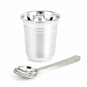 Puede incluir: Un vaso plateado pulido con bandas horizontales cerca del borde y la base, acompañado de una cuchara plateada a juego con un intrincado patrón floral en relieve a lo largo del mango y una cazoleta lisa y reflectante.