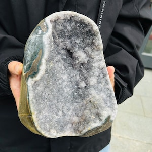 May include: A large, uncut geode with a rough, textured exterior in shades of green, brown, and gold. The interior reveals a cavity filled with sparkling, light gray and white crystals. The geode is held in a person's hands.