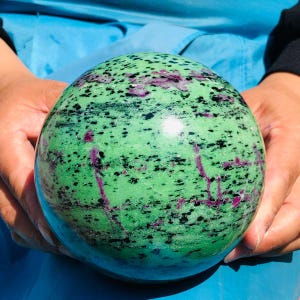 May include: A polished spherical stone featuring a vibrant green base color with scattered black mineral inclusions and distinct magenta-colored crystalline veins throughout the surface, creating a natural mottled pattern across the entire sphere.