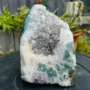 May include: A large, natural amethyst geode with a crystalline interior. The geode features shades of purple, white, and green, with a rough, textured exterior. The crystal formation is visible within the cavity, showcasing its natural beauty.