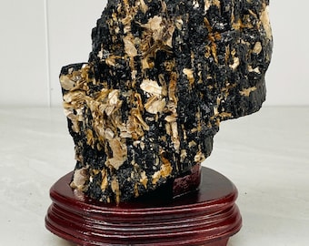 May include: A raw, jagged mineral specimen featuring a dense cluster of black tourmaline crystals interspersed with shimmering, flaky golden-brown mica deposits, mounted securely on a polished, circular dark reddish-brown wooden stand.