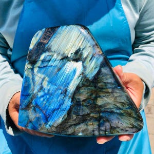 May include: A polished labradorite stone featuring a smooth, triangular shape with a vibrant iridescent blue and gold flash. The surface displays natural crystalline fractures and deep dark grey inclusions throughout the mineral specimen.