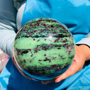 May include: A polished spherical stone featuring a vibrant green base color with prominent horizontal bands of deep magenta and black mineral inclusions. The surface is smooth and reflective, showcasing a natural pattern of contrasting colors.