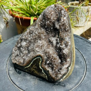 May include: A large, natural amethyst geode with a dark brown interior filled with sparkling crystals. The outer edge is a light brown with a dark green and white border. The geode is displayed on a dark surface.