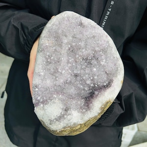 May include: A large, irregularly shaped amethyst geode. The geode features a crystalline interior with shades of purple, white, and gray. The outer edge has a rough, golden-brown appearance. The geode is held in a person's hands.