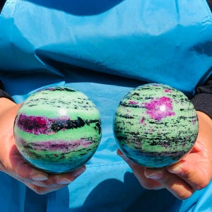 May include: Two polished spherical stones featuring a vibrant mix of light green, deep magenta, and black mineral inclusions. The spheres display a smooth, glossy surface with distinct color banding and natural patterns throughout the round forms.