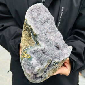 May include: A large, raw amethyst geode with a rough, textured exterior. The geode features a mix of purple, gray, and brown hues, with crystalline formations visible inside. The outer shell has areas of yellow and blue.