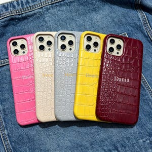 Op de afbeelding: Vijf kleurrijke telefoonhoesjes met een krokodillenleertextuur. De hoesjes zijn roze, crème, grijs, geel en bordeauxrood. Op elk hoesje staat een naam in gouden letters. De hoesjes liggen op een denim achtergrond.