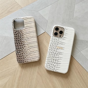 Puede incluir: Dos fundas para teléfono con textura de piel de cocodrilo. Una funda es blanquecina y marrón, la otra es blanca con un logotipo dorado. Las fundas son rectangulares con recortes para la cámara y los botones.