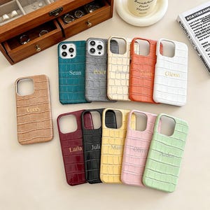 May include: A collection of phone cases in various colors, including tan, teal, gray, cream, orange, and white. Each case features a crocodile skin texture and a name in gold lettering. The cases are arranged on a light-colored surface.
