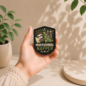 May include: A shield-shaped patch featuring a cartoon sloth sleeping under a green star-patterned blanket on a tree branch. The text PROFESSIONAL NAPPER is printed in bold cream and green letters below the sloth, with three Zs above.