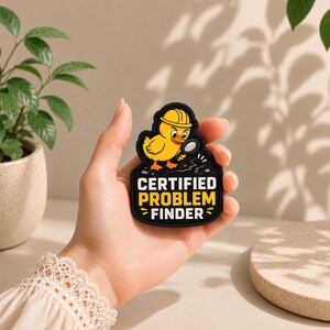 Könnte beinhalten: Ein schwarzer Gummi-Aufnäher mit einer gelben Ente, die einen Bauhelm trägt und eine Lupe über einen Riss im Boden hält. Der Text lautet CERTIFIED PROBLEM FINDER in fetten weißen und gelben Großbuchstaben.
