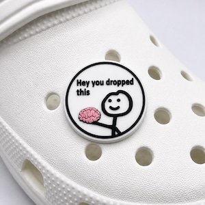 May include: A white shoe with a round, black and white charm. The charm features a stick figure holding a pink brain, with the text "Hey you dropped this".