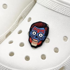 May include: A colourful shoe charm with a zombie face design. The charm has a black outline with red hair, blue skin, and a red and white mouth. It is attached to a white shoe.