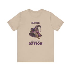 May include: A beige short-sleeve t-shirt featuring a graphic of a purple and gold witch hat with leopard print accents, feathers, and stars. The text above the hat reads SUBTLE and the text below the hat reads was never an OPTION.