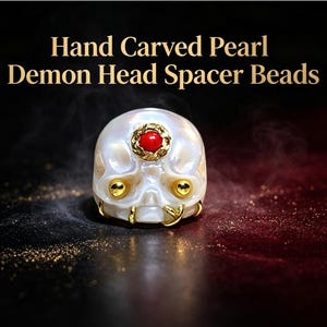 May include: A hand-carved white pearl bead shaped like a demon head, featuring a small red stone set in a gold-toned circular frame on the forehead, two gold-toned spheres for eyes, and a gold-toned hook detail on the lower jaw area.