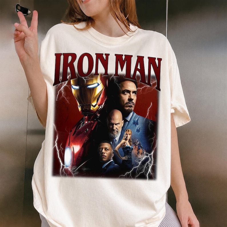 Pu&ograve; includere: T-shirt color crema con la scritta "IRON MAN" in bordeaux. La grafica presenta il casco di Iron Man e un collage di personaggi del film, su sfondo rosso con fulmini.